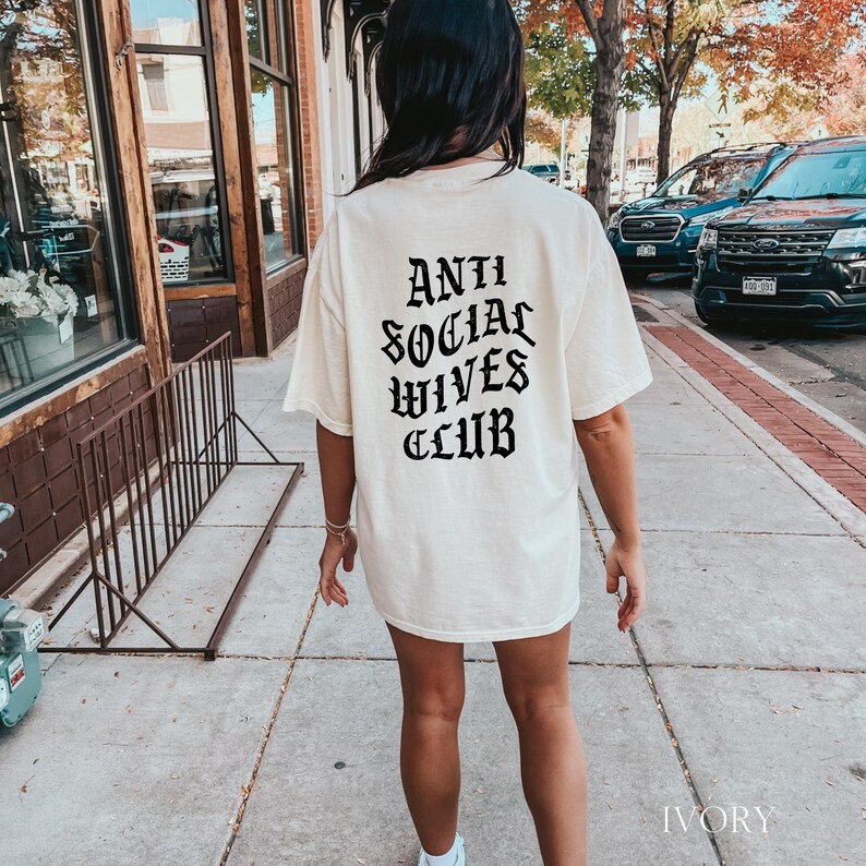 Puede incluir: Una camiseta blanca con texto negro que dice "Anti Social Wives Club" en una fuente g&oacute;tica. La camiseta la lleva puesta una persona en un entorno informal.
