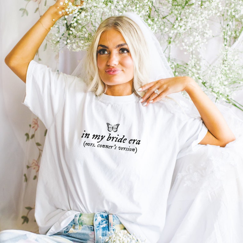 Op de afbeelding: Wit T-shirt met een vlinderprint en de tekst "in my bride era (mrs. conner's version)"