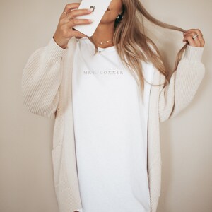 Op de afbeelding: Een wit T-shirt met lange mouwen met de tekst "MRS. CONNER" erop gedrukt. Het shirt wordt gedragen door een persoon met lang blond haar en een cr&egrave;mekleurige cardigan.