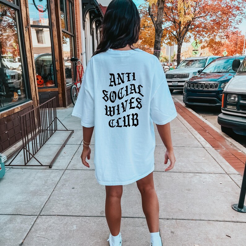 Puede incluir: Camiseta blanca de gran tama&ntilde;o con texto negro en la espalda que dice "Anti Social Wives Club" en una fuente g&oacute;tica.