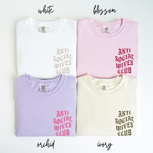 Puede incluir: Cuatro camisetas de diferentes colores con el texto "ANTI SOCIAL WIVES CLUB" en una fuente g&oacute;tica. Los colores son blanco, rosa, morado claro y crema.
