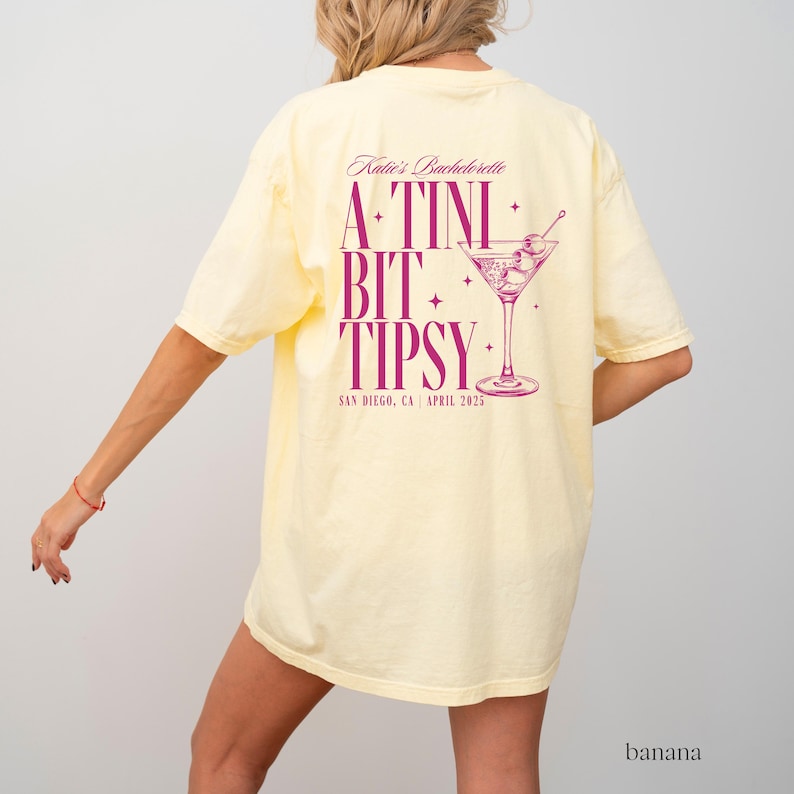 May include: A yellow t-shirt with the text "Katie's Bachelorette A-TINI BIT TIPSY SAN DIEGO, CA APRIL 2025" in pink lettering. A pink martini glass with olives is pictured above the text.