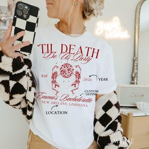 K&ouml;nnte beinhalten: Wei&szlig;es T-Shirt mit rotem Text und Grafiken. Das Design enth&auml;lt den Satz "Til Death Do Us Party", Skelette, eine Discokugel und den Text "Emma's Bachelorette, New Orleans, Louisiana, 2024."