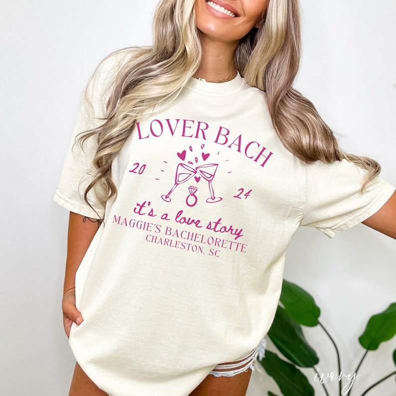 May include: A white t-shirt with the text "Lover Bach" in pink lettering. The shirt also features a pink illustration of two champagne glasses clinking, with hearts and a ring. The text "it's a love story" is below the illustration, followed by "Maggie's Bachelorette Charleston, SC".