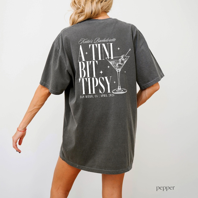 May include: A dark grey t-shirt with a white graphic that says "Katie's Bachelorette A TINI BIT TIPSY" with a martini glass illustration and the text "San Diego, CA | April 2023".