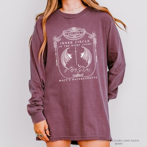 Op de afbeelding: Bessenkleurig shirt met lange mouwen en een witte afbeelding. De afbeelding bevat de tekst "INNER CIRCLE OF THE NIGHT COURT" en "MACY'S BACHELORETTE" met een ontwerp van vleugels, een zwaard, bergen en de tekst "EST 2025".