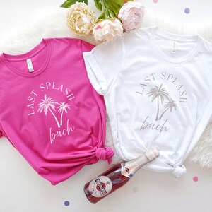 Puede incluir: Dos camisetas blancas con el texto "Last Splash Bach" y palmeras impresas en ellas. Una camiseta es rosa y la otra es blanca. Una botella de vino rosado est&aacute; en el centro de la imagen.