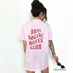 Puede incluir: Camiseta rosa con el texto "ANTI SOCIAL WIVES CLUB" en letras g&oacute;ticas rojas en la parte posterior.