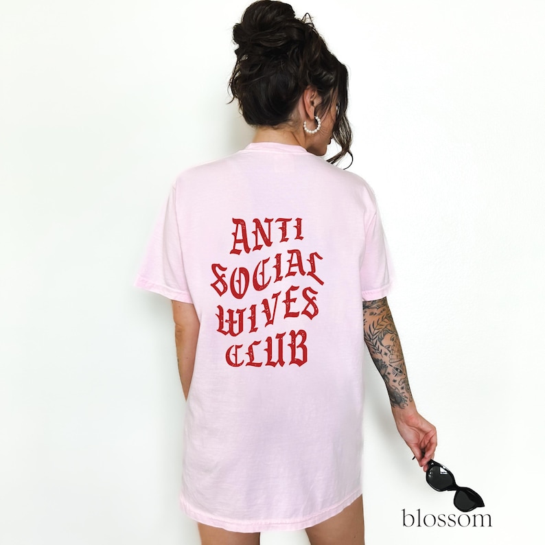 Puede incluir: Camiseta rosa con el texto "ANTI SOCIAL WIVES CLUB" en letras g&oacute;ticas rojas en la parte posterior.