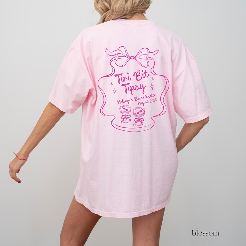 May include: Pink t-shirt with a pink graphic design that says "Tini Bit Tipsy" with a bow and stars. The text below says "Kelsey's Bachelorette August 2023".