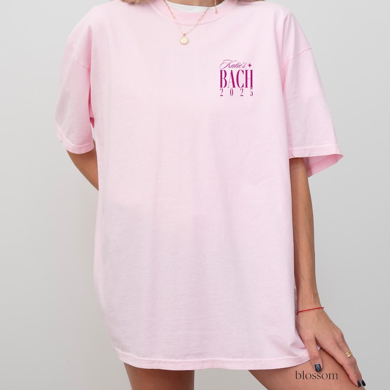 May include: Pink oversized t-shirt with the text "Katie's Bach 2023" printed on the front in purple.