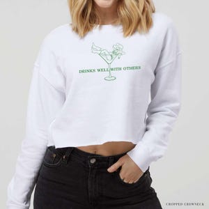 May include: White cropped sweatshirt with long sleeves featuring a green line drawing of a person lounging in a martini glass. The text "Drinks well with others" is printed below the image.