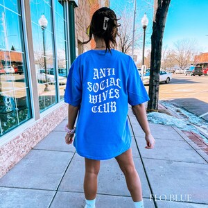 Puede incluir: Una camiseta azul real con el texto "ANTI SOCIAL WIVES CLUB" impreso en blanco en la parte posterior. El texto est&aacute; en una fuente g&oacute;tica.