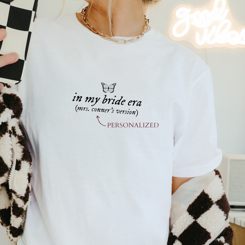 Op de afbeelding: Wit T-shirt met de tekst "in my bride era (mrs. conner's version)" en een vlindergrafiek. De tekst "PERSONALIZED" staat onder de tekst.