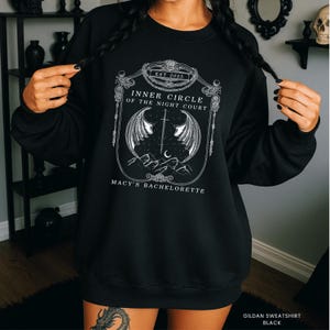 Op de afbeelding: Zwart sweatshirt met een wit grafisch ontwerp. Het ontwerp bevat de tekst "Inner Circle of the Night Court" en "Macy's Bachelorette" in een sierlijke lijst, samen met een zwaard, vleugels, bergen en een halve maan.