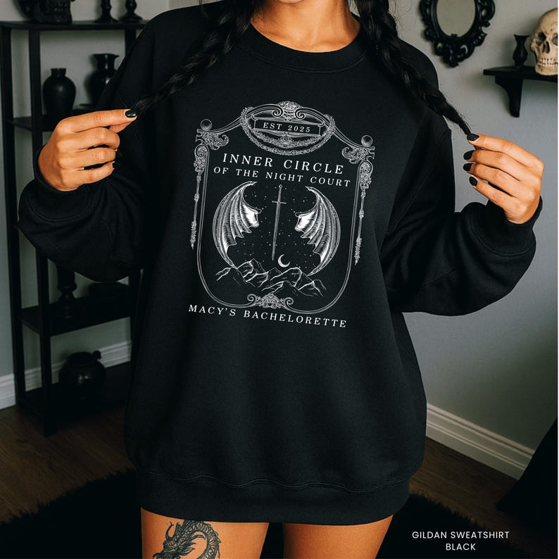 Op de afbeelding: Zwart sweatshirt met een wit grafisch ontwerp. Het ontwerp bevat de tekst "Inner Circle of the Night Court" en "Macy's Bachelorette" in een sierlijke lijst, samen met een zwaard, vleugels, bergen en een halve maan.