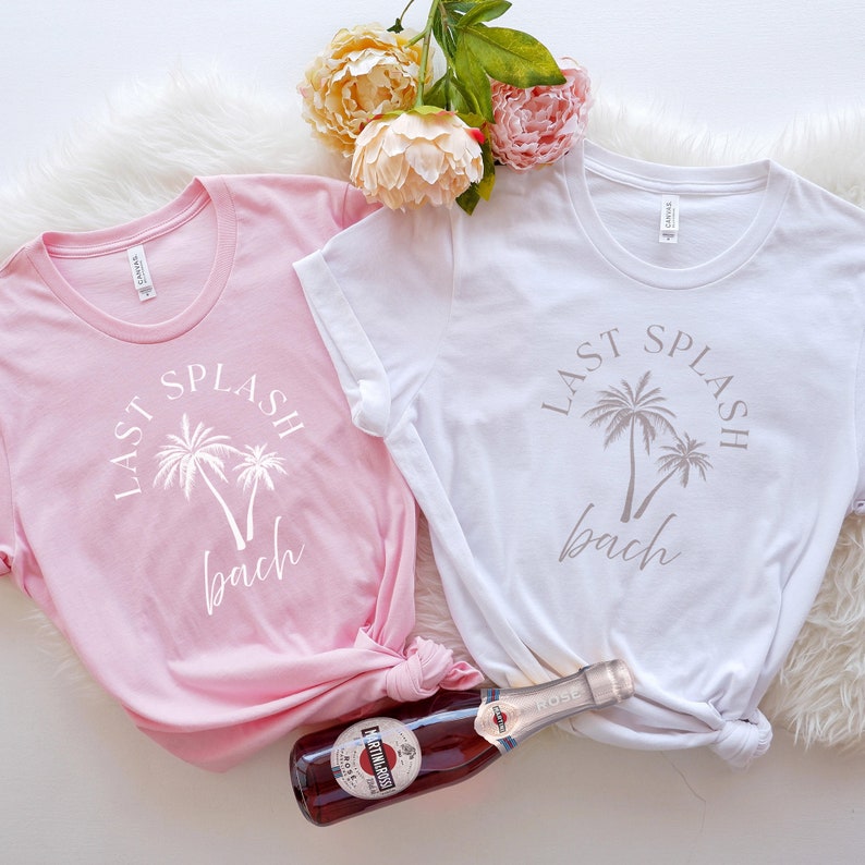 Puede incluir: Dos camisetas blancas con el texto "Last Splash Bach" y gr&aacute;ficos de palmeras. Una camiseta es rosa y la otra es blanca. Una botella de vino rosado est&aacute; en primer plano.