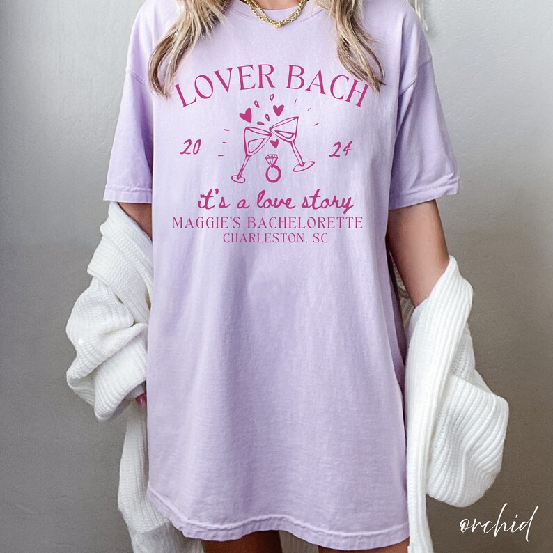 May include: A lavender-colored t-shirt with the text "Lover Bach" in pink lettering. The shirt also features a pink illustration of two champagne flutes with a diamond ring in the middle. The text "it's a love story" is below the illustration, followed by "Maggie's Bachelorette Charleston, SC".