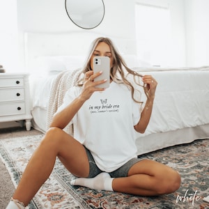 Op de afbeelding: Een wit T-shirt met de tekst "in my bride era (mrs. conner's version)" en een vlindergrafiek.