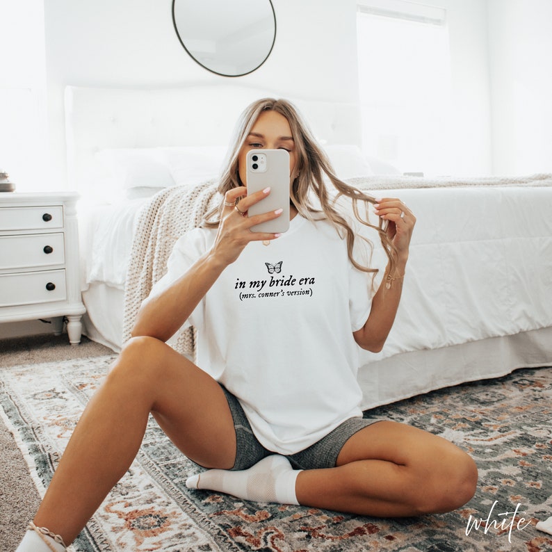 Op de afbeelding: Een wit T-shirt met de tekst "in my bride era (mrs. conner's version)" en een vlindergrafiek.