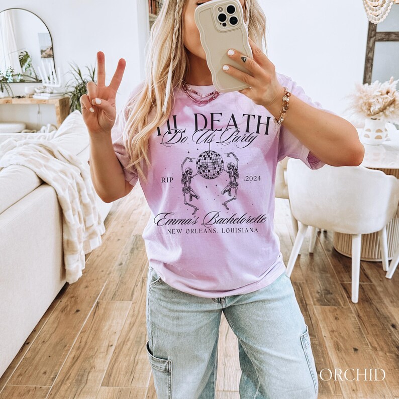 K&ouml;nnte beinhalten: Helllila T-Shirt mit dem Text "Til Death Do Us Party" und "Emma's Bachelorette New Orleans, Louisiana". Das Shirt zeigt eine Grafik von zwei Skeletten, die um eine Discokugel tanzen. Das Jahr "2024" ist ebenfalls auf dem Shirt aufgedruckt.