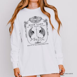 Op de afbeelding: Wit shirt met lange mouwen met een grafisch ontwerp. Het ontwerp bevat de tekst "EST 2025", "HIGH LADY OF THE NIGHT COURT" en "MACY'S BACHELORETTE". De afbeelding toont vleugels, een zwaard en een maan.