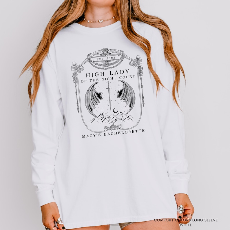 Op de afbeelding: Wit shirt met lange mouwen met een grafisch ontwerp. Het ontwerp bevat de tekst "EST 2025", "HIGH LADY OF THE NIGHT COURT" en "MACY'S BACHELORETTE". De afbeelding toont vleugels, een zwaard en een maan.