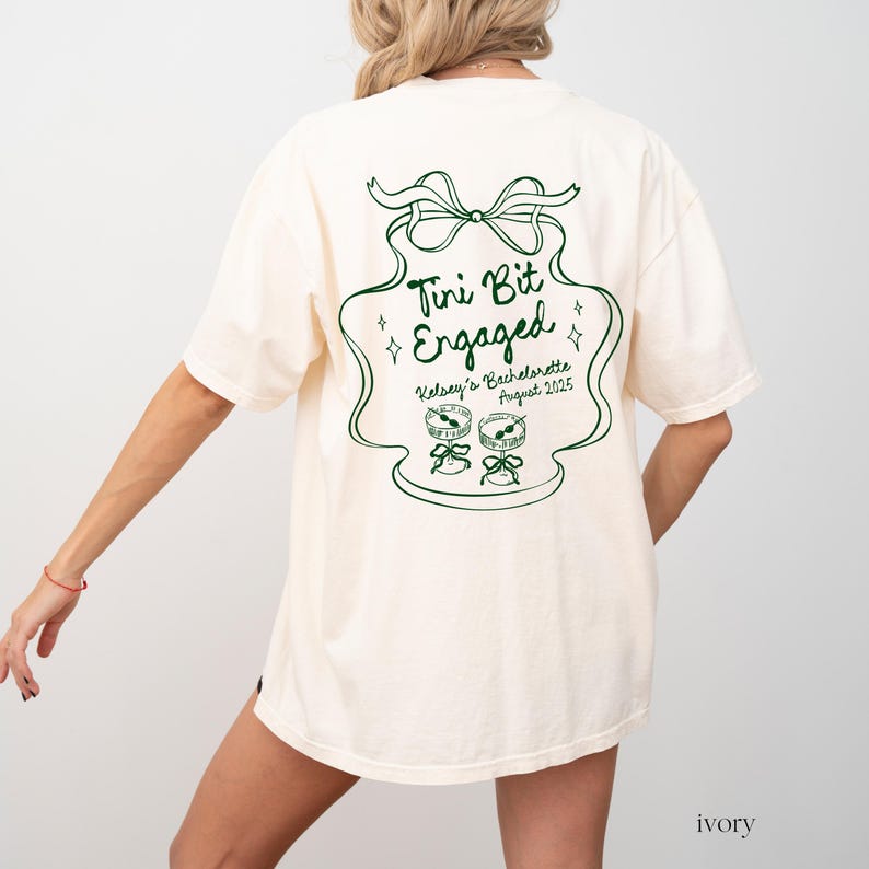 May include: Ivory t-shirt with a green graphic that says "Tini Bit Engaged" with a bow and two martini glasses. The text below the graphic says "Kelsey's Bachelorette August 2025".