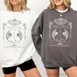 Op de afbeelding: Twee sweatshirts, &eacute;&eacute;n witte en &eacute;&eacute;n houtskoolkleurige, met een gedetailleerd grafisch ontwerp. Het witte sweatshirt toont "HIGH LADY OF THE NIGHT COURT" en de houtskoolkleurige leest "INNER CIRCLE OF THE NIGHT COURT". Beide bevatten "EST 2025" en "MACY'S BACHELORETTE".