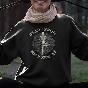 Op de afbeelding: Een zwart sweatshirt met een witte grafische print van een skelet dat yoga doet in een mandala. De tekst op het sweatshirt luidt "Dead Inside But Zen AF".