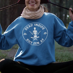 Peut inclure: Un sweat-shirt bleu avec un graphique blanc d'un squelette assis en position de méditation avec le texte "Dead Inside But Zen AF" dans un cercle autour du squelette.