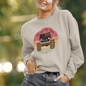 May include: A light beige sweatshirt featuring a vintage-style graphic of a jeep with an American flag on the bonnet. The graphic is surrounded by a red, orange and yellow sunset and the text "Vintage Off Road".