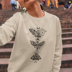 May include: A beige sweatshirt with a black and white graphic print of three moths and geometric shapes. The moths are arranged vertically with a crescent moon at the top and bottom of the design.
