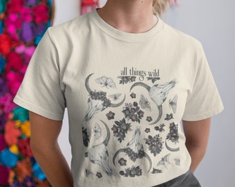 Può includere: Una t-shirt color crema con una stampa grafica di uno scheletro con fiori, farfalle e lune crescenti. Il testo "all things wild" è stampato sulla t-shirt.