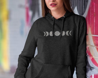 Puede incluir: Una sudadera con capucha negra con un gráfico de las fases de la luna en diferentes etapas. El gráfico es de color gris claro con un sutil efecto brillante. La sudadera la lleva puesta una persona de pie contra una pared de ladrillo.