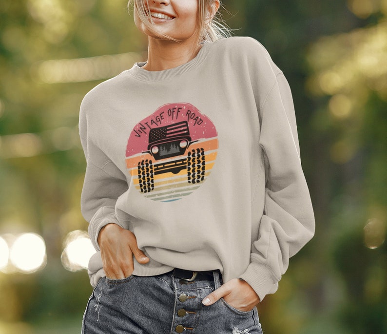 Puede incluir: Una sudadera de color beige claro con un gr&aacute;fico de estilo vintage de un jeep con una bandera estadounidense en el cap&oacute;. El gr&aacute;fico est&aacute; rodeado por una puesta de sol roja, naranja y amarilla y el texto "Vintage Off Road".
