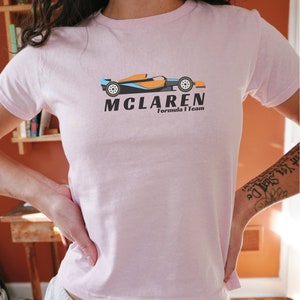 Puede incluir: Una camiseta rosa con un logotipo negro y naranja de McLaren Formula 1 Team que presenta un coche de carreras de dibujos animados.