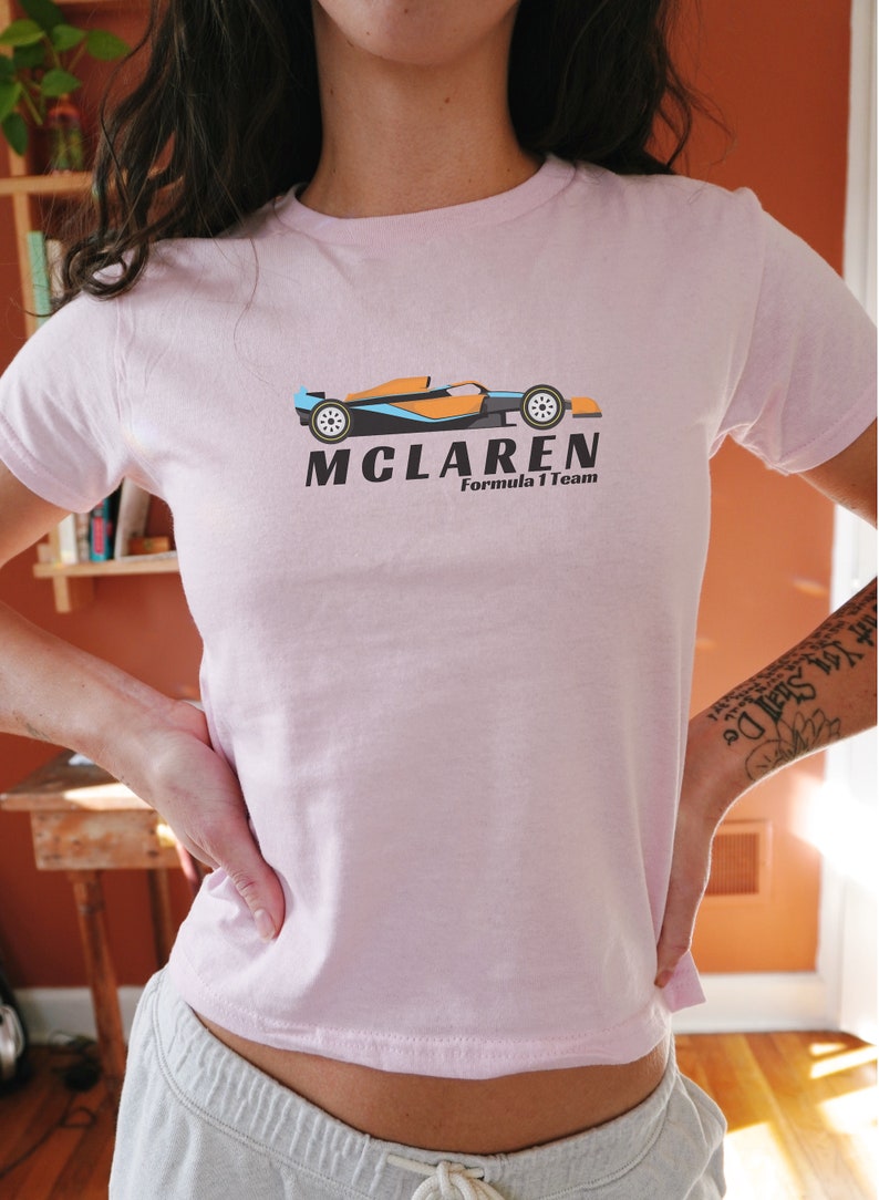 Puede incluir: Una camiseta rosa con un logotipo negro y naranja de McLaren Formula 1 Team que presenta un coche de carreras de dibujos animados.
