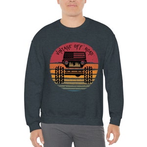 Puede incluir: Una sudadera azul oscuro con un gr&aacute;fico de un jeep vintage todoterreno. El gr&aacute;fico presenta una silueta de un jeep con una bandera estadounidense en el cap&oacute;, sobre un fondo de puesta de sol con un esquema de color retro. El texto "Vintage Off Road" est&aacute; escrito en un arco circular alrededor del jeep.