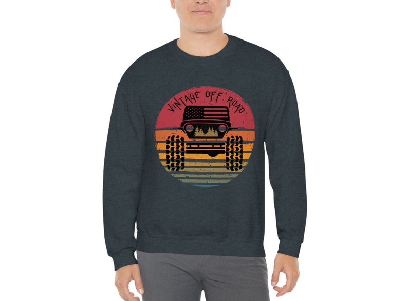 Puede incluir: Una sudadera azul oscuro con un gr&aacute;fico de un jeep vintage todoterreno. El gr&aacute;fico presenta una silueta de un jeep con una bandera estadounidense en el cap&oacute;, sobre un fondo de puesta de sol con un esquema de color retro. El texto "Vintage Off Road" est&aacute; escrito en un arco circular alrededor del jeep.