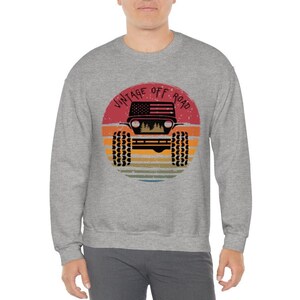 Puede incluir: Una sudadera gris con un gr&aacute;fico de un jeep vintage todoterreno. El gr&aacute;fico presenta una bandera estadounidense roja, blanca y azul, una puesta de sol y el texto "Vintage Off Road".