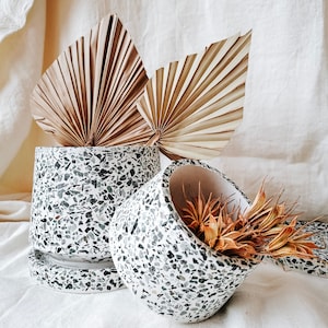 Op de afbeelding: Twee witte keramische potten met een zwart-wit gespikkeld patroon. De potten staan op een wit stoffen oppervlak. Gedroogde palmbladeren zijn op de achtergrond.