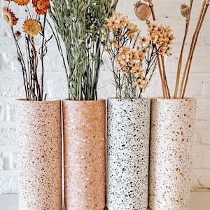 Op de afbeelding: Vier terrazzo vazen in verschillende kleuren, gevuld met gedroogde bloemen en grassen. De vazen zijn allemaal even groot en hebben dezelfde vorm, en ze zijn in een rij op een wit oppervlak gerangschikt.