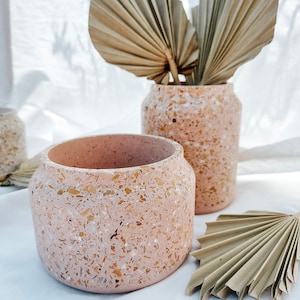Può includere: Due vasi in terrazzo rosa con una finitura macchiettata. Il vaso più grande è più alto e più stretto del più piccolo. Entrambi i vasi sono su una superficie bianca con foglie di palma secche.