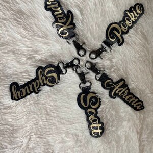 Puede incluir: Cuatro llaveros de cuero negro con letras doradas. Los nombres en los llaveros son Kendra, Pookie, Adrena y Scott.