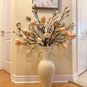 May include: A white ceramic vase with a wide mouth and two handles holds a bouquet of peach-colored magnolia blossoms.