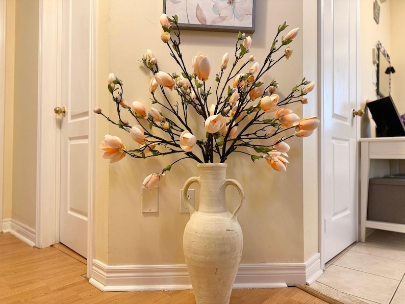 May include: A white ceramic vase with a wide mouth and two handles holds a bouquet of peach-colored magnolia blossoms.