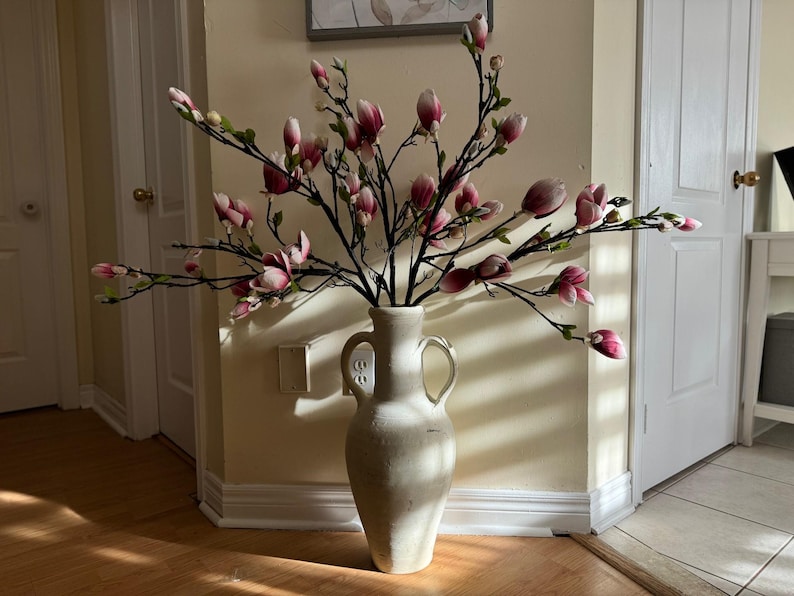 May include: A white ceramic vase with two handles, filled with a large arrangement of pink and white magnolia blossoms.
