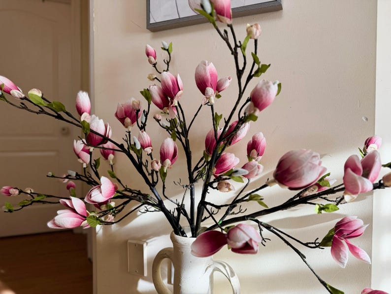 May include: A bouquet of pink and white magnolia flowers in a white vase. The flowers are arranged in a vase with a rustic, farmhouse style.
