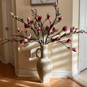 May include: A white ceramic vase with two handles, filled with a bouquet of pink and white magnolia flowers.